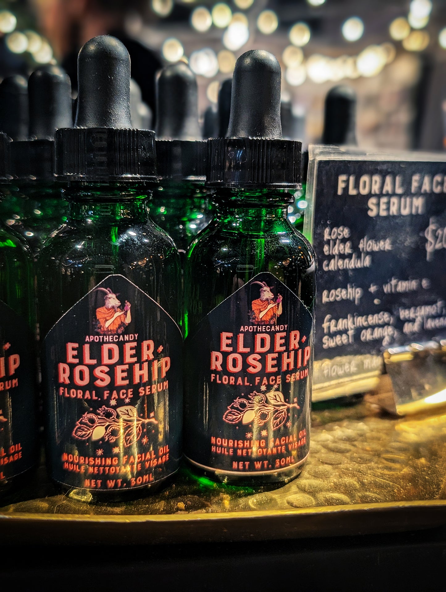 Green glass dropper top bottles of Elder and Rosehip Floral Face Serum are displayed on a golden tray with twinkling lights in the background