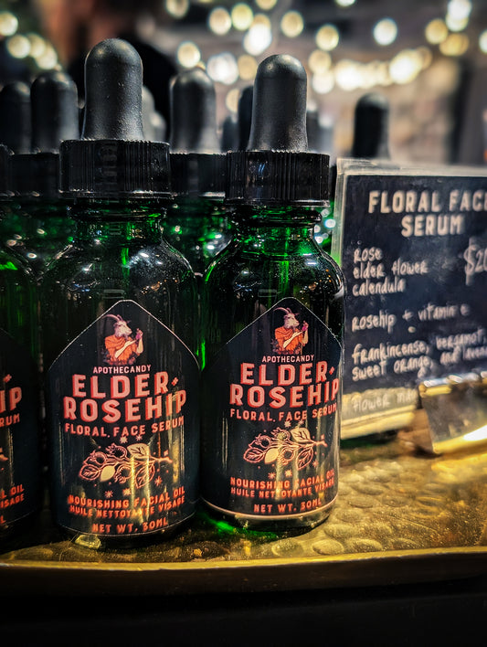 Green glass dropper top bottles of Elder and Rosehip Floral Face Serum are displayed on a golden tray with twinkling lights in the background