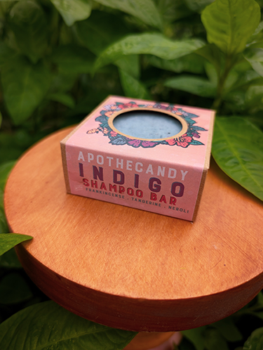 Apothecandy Indigo shampoo bar in a blush box on a wooden surface with verdant green indigo leaves in the background