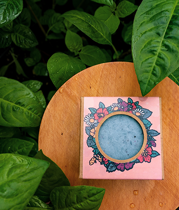 An Indigo Shampoo bar in a blush coloured box sits atop a wooden stand surrounded by verdant green indigo leaves